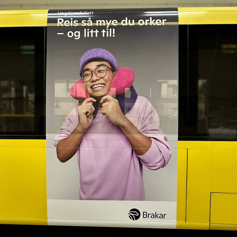 Nærbilde av siden på en gul buss med stående reklamebudskap for Brakar. En ung person i violett genser og briller som smiler med en nakkepute.