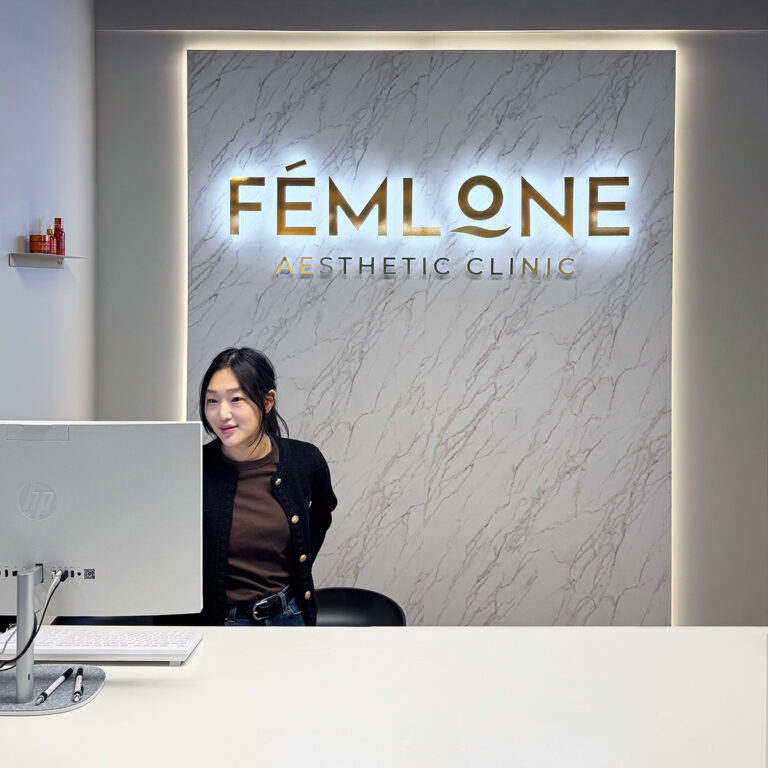 Kvinne foran stort LED-lysskilt innvendig i en skjønnhetssalong. LED-skilt bakbelyst med marmorfoliert front og bakbelyste bokstaver i gull for logoen Femlone Aesthetic Clinic
