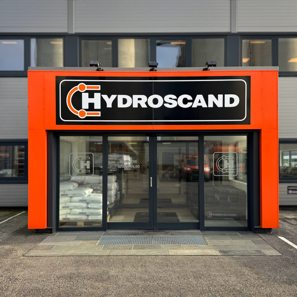 Inngansparti med skyvedører omkranset av en oransje portal i folierte plater og stort sort plateskilt for Hydroscand