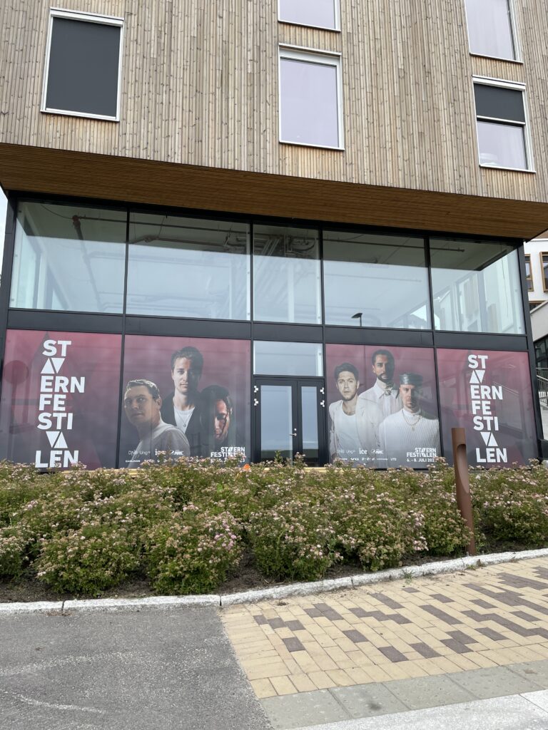 Moderne bygning med lysegrå trepanel og store vinduer. Nedre del av fasaden har røde plakater med teksten "STAVERNFESTIVALEN" og portretter av mennesker. Hvite blomster i hagen foran bygningen