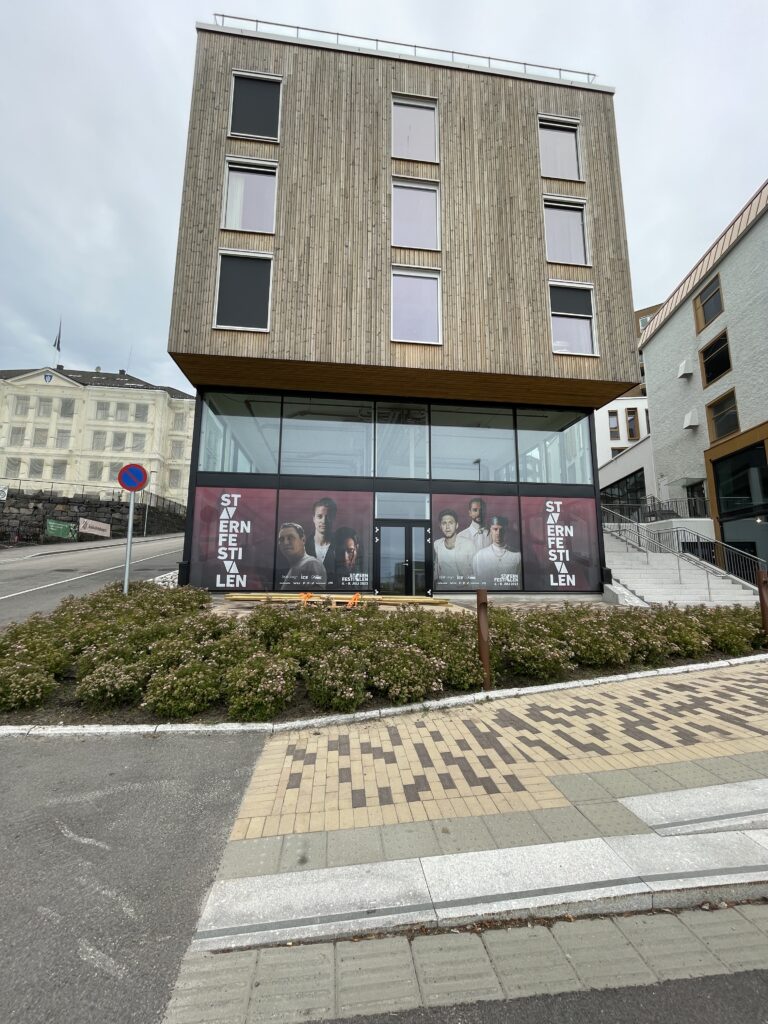 Moderne bygning med tre etasjer og vertikale trelameller. Glassfasade i første etasje med vindusdekor som viser "Starvernfesitvalen"-tekst og portretter av mennesker