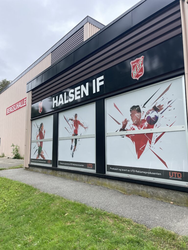 Nærbilde av plateskilt Halsen IF på Bergslihallen med folierte vinduer av håndballspillere. Arenaskilt på en idrettshall.