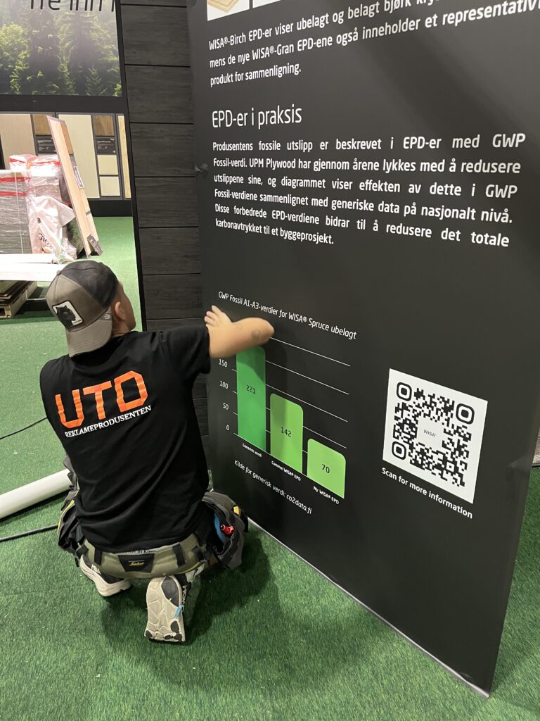 Person i UTD-jakke monterer interaktiv utstilling om EPD-er og GWP-verdier for byggeprodukter, med grønt stolpediagram og QR-kode