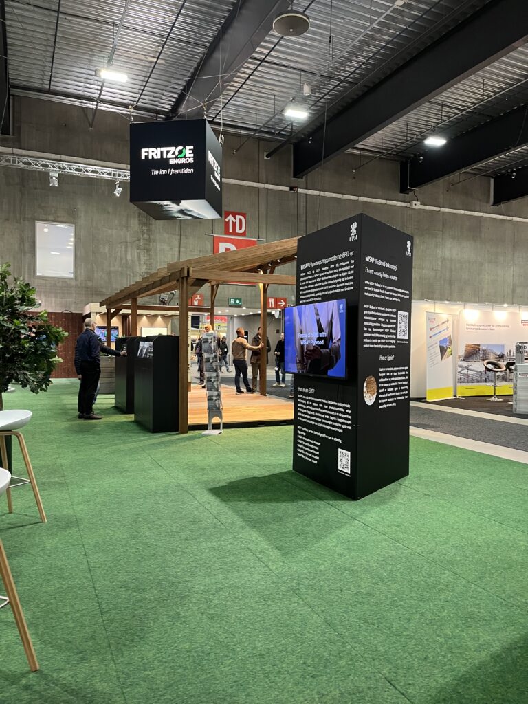 Utstillingspaviljong med trestruktur og svarte informasjonspaneler på grønt gulv i stor industriell hall. Fritze Energos logo henger fra taket, og besøkende utforsker utstillingen