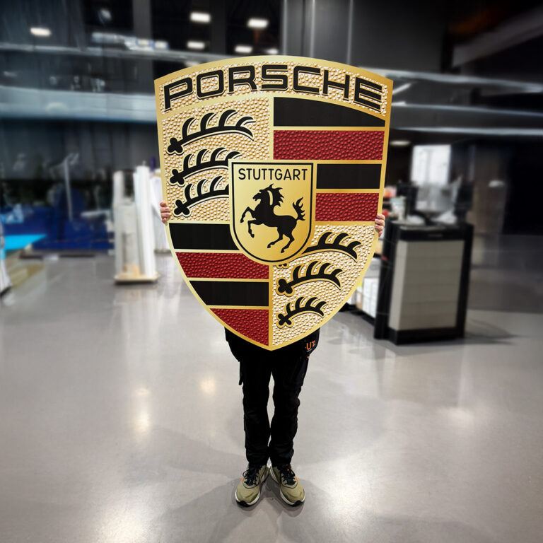 Person som holder opp et stort utfrest skilt av Porsche logo foran seg.
