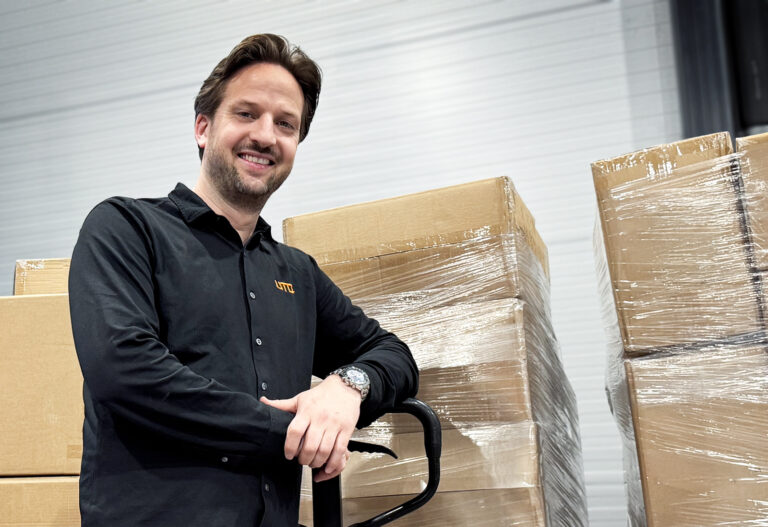 Mann smilende i sort profilskjorte med UTD logo foran esker på pall pakket inn i plast