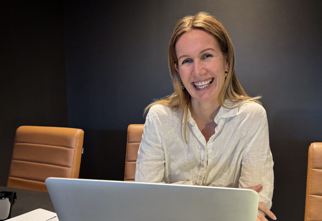 Kvinne smilende i hvit skjorte foran laptop på et møterom
