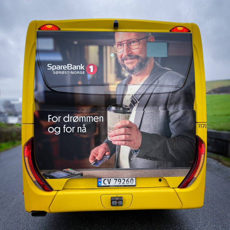 Baksiden av en gul buss sett i trafikken med helfoliert bakside for Sparebank 1 Sørøst-Norge. Bussreklame