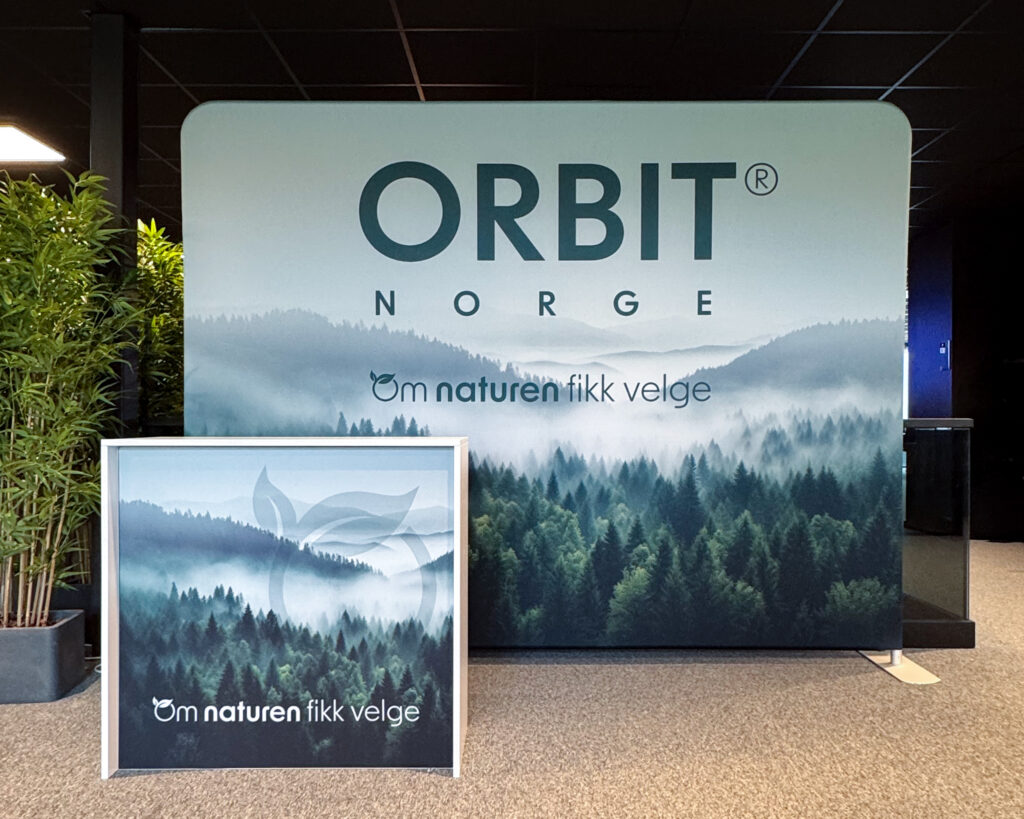 Orbit Norge-utstilling med banner og plakat som viser skoglandskap og slagordet "Om naturen fikk velge". Messevegg med bord.