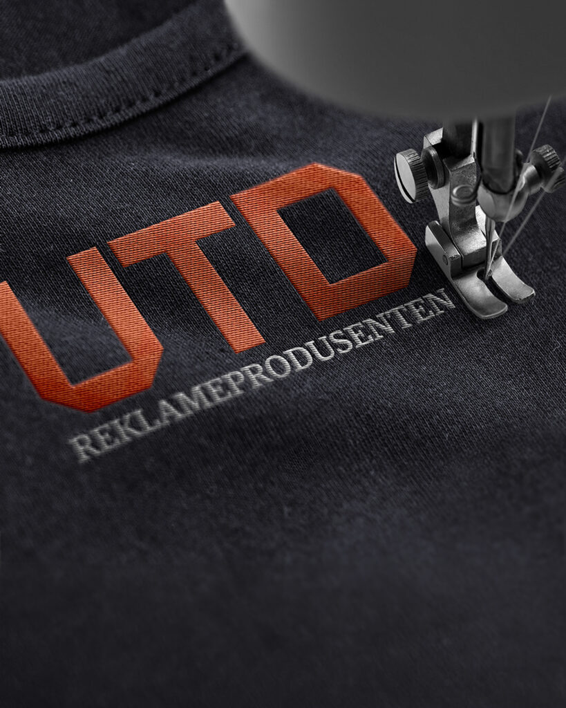 UTO-logo med tekst "REKLAMEPRODUSENTEN" brodert på mørk stoff med sytråd og nål