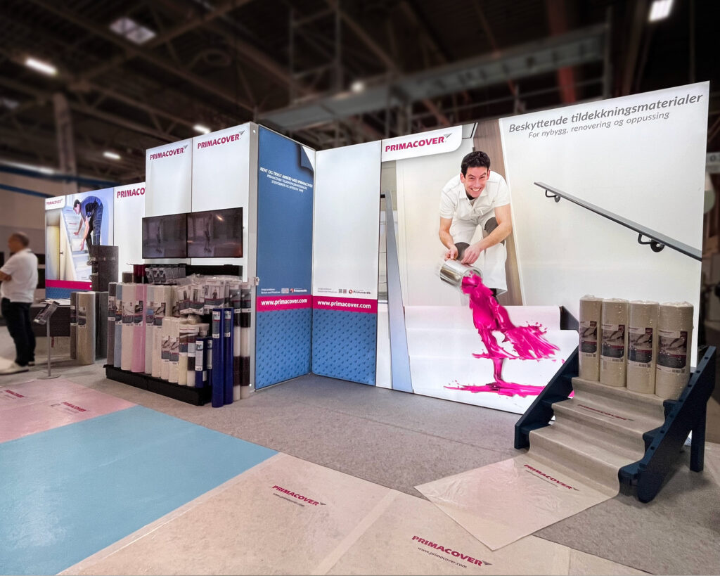 Primaverde-messestand på byggmesse med produktdisplay, fargekart og demonstrasjon av malingapplikasjon på vegg. Store messevegger med bakbelyst tekstilduk.