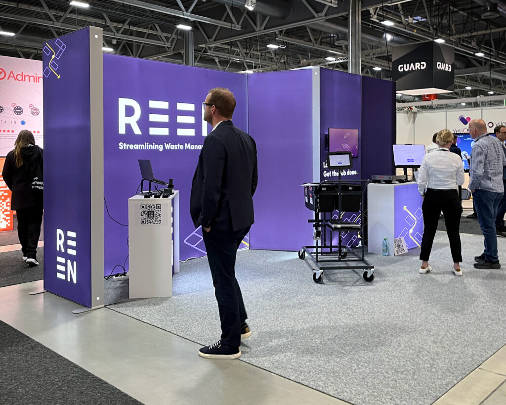 REEN-utstilling på messe med lilla vegger, besøkende som utforsker avfallshåndteringsteknologi, og interaktive skjermer i en åpen messestand.