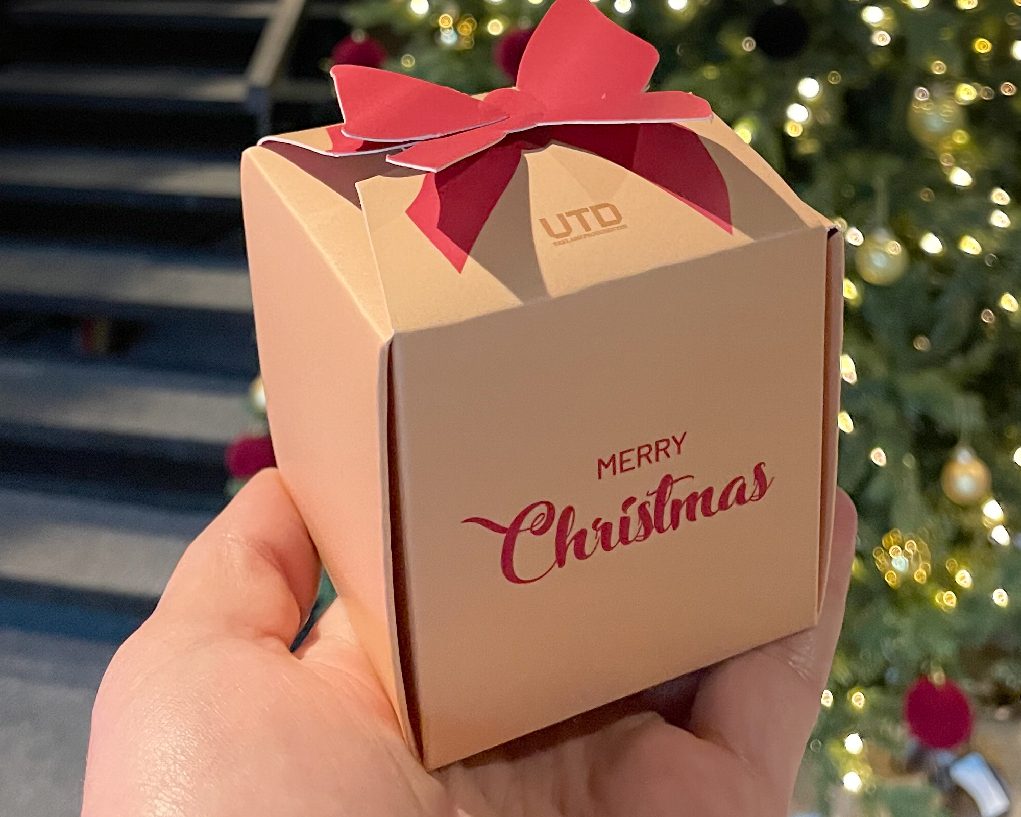 Gaveeske med rød sløyfe og teksten "Merry Christmas" foran et juletre i bakgrunnen.