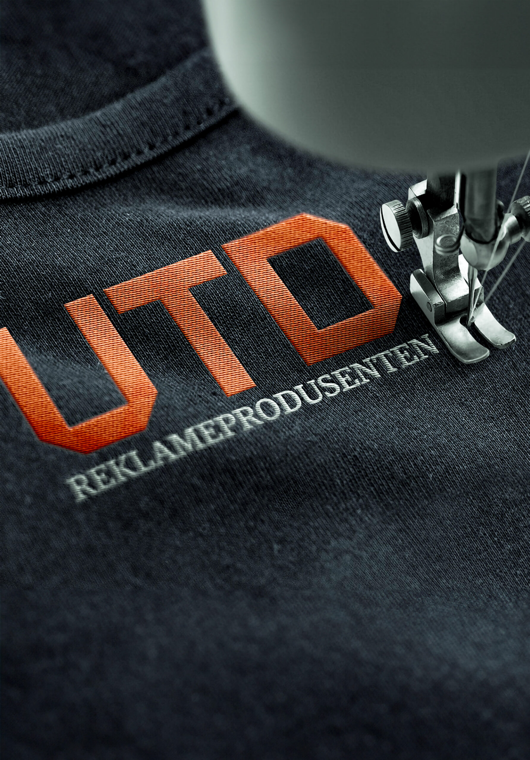 Klær med oransjeopp-brodert UTD-logo og teksten "REKLAMEPRODUSENTEN" med en sy-maskinnål i fokus