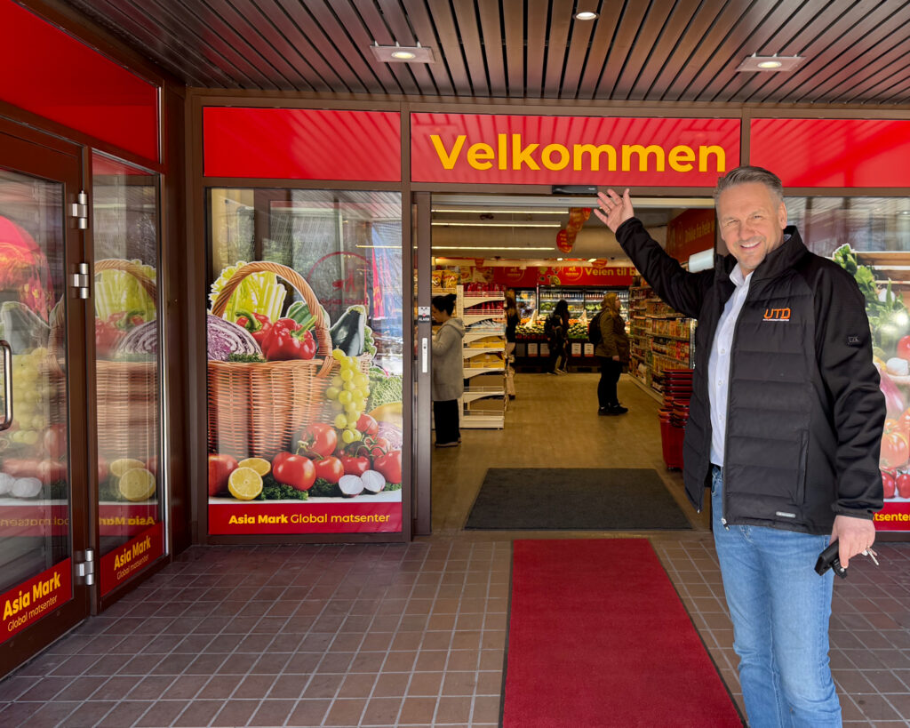 Mann i UTD-jakke ved Asia Mark-inngang med «Velkommen»-skilt og vindusdekor — prosjektansvarlig fra UTD ved ferdigstilt butikk