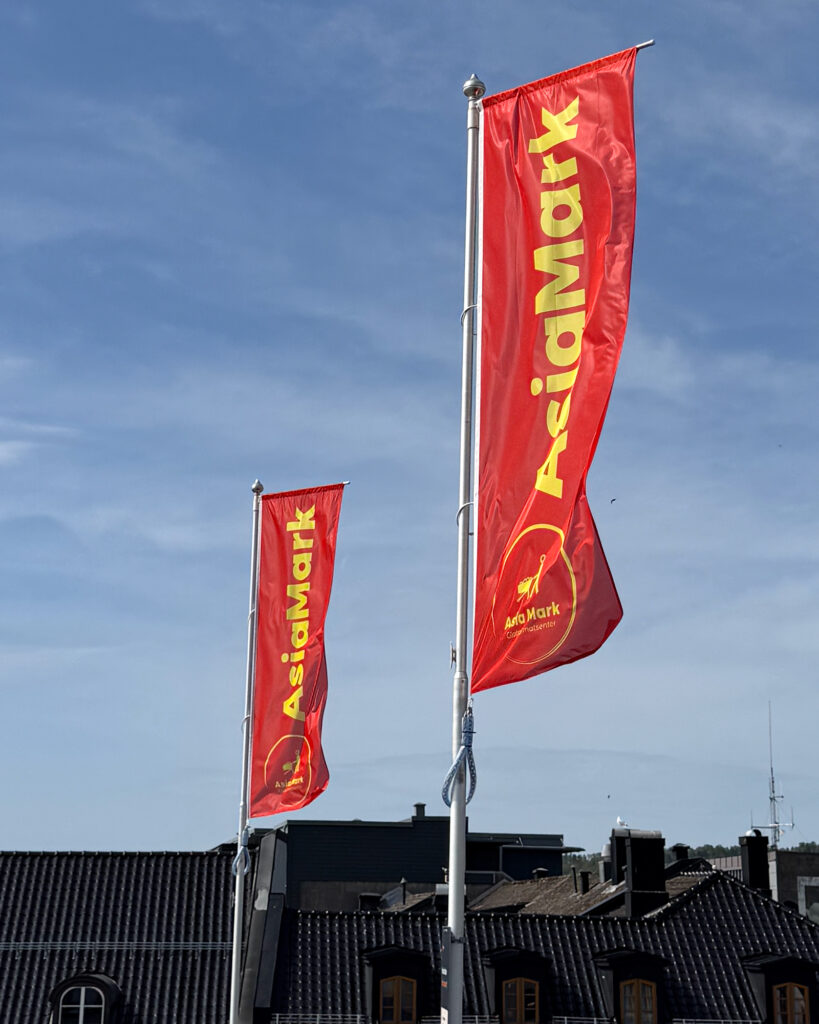 To røde Asia Mark-flagg med gul logo vaier i vinden mot blå himmel — flaggproduksjon og profilering levert av UTD